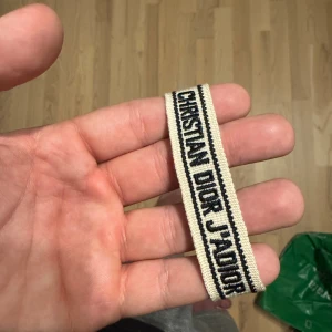 Christian Dior J'Adior armband - Snyggt vävt armband från Christian Dior med texten 'J'Adior' i svart på beige bakgrund. Armbandet har svarta ränder längs kanterna och avslutas med flätade snören och tofsar. Perfekt accessoar för dig som gillar designer och vill sticka ut.