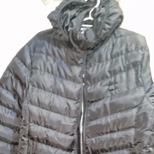 Svart pufferjacka med värmefunktion - Svart pufferjacka med snygg glansig yta och horisontella quiltade sektioner. Jackan har huva och dragkedja framtill. På bröstet sitter en knapp för värmefunktion, perfekt när det är kallt ute. Materialet är syntet och jackan har en normal passform.