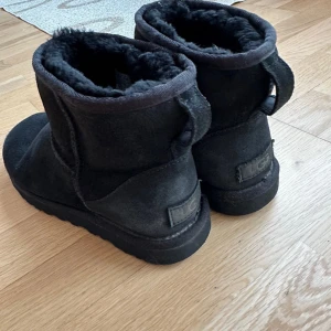 Svarta UGG boots i mocka storlek 38 - Mysiga svarta UGG boots i mjuk mocka med fluffigt foder och platt sula. Klassisk rund tå och enkel design som passar perfekt till kalla dagar. UGG-logga bak på hälen och dragflik för enkel påtagning.