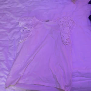 Vit blus med broderade volangärmar - Supersöt vit blus med broderade volangärmar och hålmönster. Ärmlös modell med romantiska detaljer och rund halsringning. Perfekt för dig som gillar en feminin och fräsch stil.