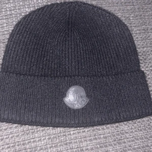 Svart Moncler stickad mössa - Snygg svart mössa från Moncler i ribbstickad ullblandning. Klassisk beanie-modell med uppvikt kant och Moncler-logga framtill i svart. Perfekt för dig som vill ha en stilren och lyxig look under kyliga dagar.