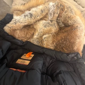 Svart Parajumpers dunjacka med päls - Säljer en svart Parajumpers dunjacka med fluffig naturfärgad päls runt huvan. Jackan har coola detaljer som orange rem och flera snygga märken på insidan. Perfekt för dig som vill ha en varm och stilren vinterjacka med exklusiv känsla.