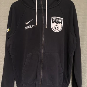 Svart Nike hoodie Qviding FIF M - Svart hoodie från Nike med Qviding FIF-logga och Stadium-tryck på bröstet. Mjuk insida, perfekt för fotboll eller vardags. Mycket fint skick. Längd: ca 63cm, Bredd: ca 56cm