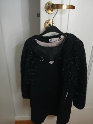 Svart glittrig kortklänning H&M - Svart kortklänning från H&M med glittrig, fluffig överdel och långärmade ärmar. Framsidan har ett sött ansikte med broderad nos och ögon, samt ljusrosa detaljer vid halsen. Perfekt för dig som gillar unika och lekfulla plagg.