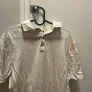 Vit kortärmad henley med krage - Säljer en vit henley-skjorta med klassisk krage och två knappar framtill. Skjortan har korta ärmar och är gjord i mjukt bomullsmaterial, perfekt för en chill och clean look. Enkel att matcha med jeans eller shorts.