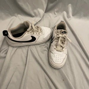 Vita Nike sneakers med svart swoosh - Säljer ett par fräscha vita Nike sneakers med klassisk svart swoosh på sidan. Skorna har snörning, perforerad tå och rund sula. Tillverkade i syntet och skinn med bekväm mesh på insidan. Perfekt för dig som gillar stilrena och sportiga sneakers.