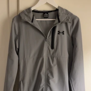 Grå vindjacka från Under Armour - Snygg grå vindjacka från Under Armour med huva och svart logga på bröstet. Jackan har dragkedja framtill och en extra ficka med dragkedja på bröstet. Perfekt för aktiva dagar och har en clean, sportig look.