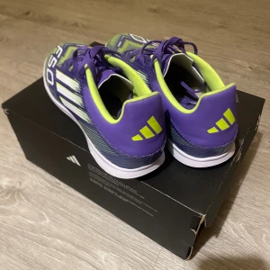 Adidas F50 lila fotbollsskor - Säljer ett par Adidas F50 fotbollsskor i lila med neon-gula och vita detaljer. Skorna har snörning, platt sula och ett grafiskt mönster med F50-tryck på ovansidan. Insidan är neon-gul och yttermaterialet är syntet. Perfekt för dig som vill sticka ut på planen.