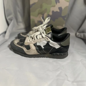 Valentino Rockrunner  - Snygga Valentino Rockrunner sneakers med grå, svarta och beige camouflagepaneler i mocka och skinn. Platt sula med nitar på hälen, rund tå och snörning framtill. Ikonisk logga på plösen och cool armykänsla. Perfekt för dig som gillar streetstyle och designer.