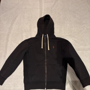 Svart hoodie från Polo Ralph Lauren - Svart zip hoodie från Polo Ralph Lauren med dragkedja och vit snörning i huvan. Klassisk röd broderad logga på bröstet och två fickor framtill. Använd men i bra skick, inga missfärgningar eller andra deffekter. Passformen är lite liten Nypris 2395 Mittpris 495