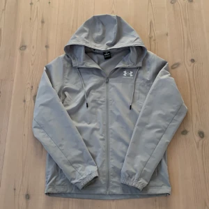 Grå vindjacka från Under Armour - Snygg grå vindjacka från Under Armour med huva och dragsko. Jackan har dragkedja framtill, elastiska muddar och meshfoder på insidan. Perfekt för dig som gillar sportig stil och vill ha något lätt och funktionellt.