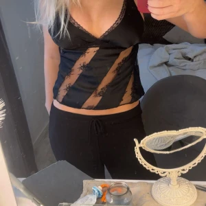 Svart satinlinnetopp med spetsdetaljer - Snygg svart linnetopp i satin med transparenta spetsinslag och v-ringning. Toppen har smala axelband och dekorativa spetskanter längs urringningen samt diagonala spetsränder framtill. Perfekt för dig som gillar en trendig och feminin look.