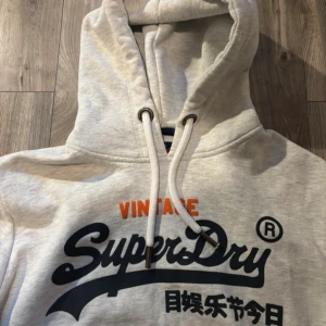 Grå hoodie från Superdry med tryck - Säljer en ljusgrå hoodie från Superdry med stort, coolt tryck i marinblått och orange på bröstet. Tröjan har huva med snörning, ribbade muddar och en klassisk känguruficka framtill. Perfekt för dig som gillar streetwear och snygga detaljer.
