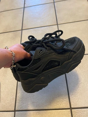 Svarta chunky sneakers med plattform - Chunky svarta sneakers med grov plattformssula och snörning. Ovandelen har meshdetaljer och syntetpaneler som ger en cool streetstil. Skorna har rund tå och tjocka svarta skosnören. Perfekt för dig som gillar att sticka ut med dina skor.