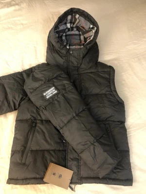 Svart dunväst från Burberry - Säljer en svart dunväst från Burberry med avtagbara ärmar och huva. Västen har quiltad design, dragkedja och tryckknappar framtill samt två fickor med dragkedja. Insidan av huvan har klassiskt rutigt Burberry-mönster i grått, vitt, svart och rött. Stor logopatch på ena ärmen. Ev byte