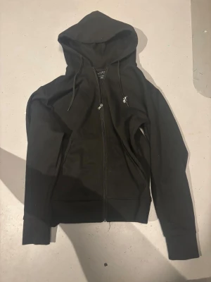 Svart hoodie från Polo Ralph Lauren - Svart hoodie från Polo Ralph Lauren med klassisk logga broderad på bröstet. Tröjan har huva med snörning, dragkedja framtill och två fickor. Materialet är mjuk bomull och modellen är normal passform med långa ärmar. Perfekt för chill dagar.