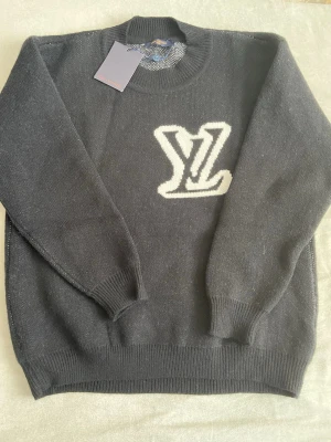 Svart stickad tröja Louis Vuitton - Snygg svart stickad tröja från Louis Vuitton med stort vitt LV-logo på bröstet. Tröjan har ribbade muddar vid hals, ärmslut och nederkant. Perfekt för dig som vill ha en exklusiv och stilren look med premiumkänsla.