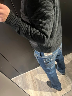 Blå True Religion jeans med kontrastsömmar - Säljer ett par blå jeans från True Religion med ikoniska kontrastsömmar och detaljerade bakfickor med lock och knapp. Klassisk femficksmodell och normal passform. Jeansen har en snygg tvätt och coola metalldetaljer på fickorna.