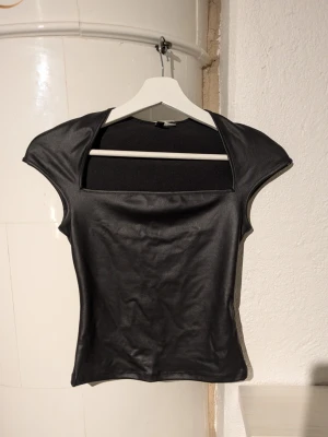 Svart croppad topp med öppen rygg - Snygg svart croppad topp med cool öppen rygg och breda axlar. Materialet har en lätt glansig finish som ger en edgy vibe. Perfekt för att sticka ut med en unik siluett och modern look.