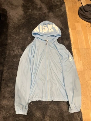 Ljusblå vindjacka från Risky - Snygg ljusblå vindjacka från Risky med coolt vitt tryck på huvan och ärmen. Jackan har dragkedja framtill, huva och elastiska muddar vid ärmslut och nederkant. Perfekt för dig som gillar streetwear och vill sticka ut med en unik look.