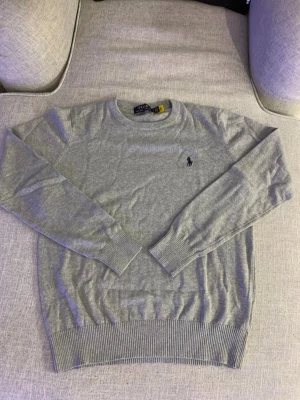 Grå stickad tröja från Polo Ralph Lauren - Snygg grå stickad tröja från Polo Ralph Lauren med klassisk rund hals och diskret broderad logga på bröstet. Tröjan har långa ärmar och ribbade muddar vid ärmslut och nederkant. Perfekt för dig som gillar stilrena och tidlösa plagg.