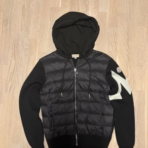 Svart cardigan  med huva från Moncler - Svart pufferjacka från Moncler med stickade ärmar och stor huva. Jackan har dragkedja framtill, två fickor och en vit blixtdetalj på ena ärmen. Materialmix av polyester och stickat ger en unik look. Perfekt för dig som vill ha något stilrent men ändå sticker ut. Kvitto finns! 