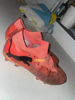 Puma fotbollsskor i orange och gul - Säljer ett par snygga fotbollsskor från Puma i en cool orange färg med gula detaljer och svarta Puma-loggor på sidorna. Skorna har platt sula med dobbar för gräsplan och en strumpliknande, stickad ovandel för skön passform. Perfekt för dig som vill sticka ut på planen!