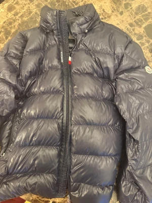 Mörkblå Moncler pufferjacka - Säljer en mörkblå pufferjacka från Moncler med hög krage och dragkedja framtill. Jackan har ett glansigt yttermaterial och klassisk Moncler-logga på ärmen. Insidan har ett coolt serietidningsmotiv och flera innerfickor. Perfekt för kalla dagar.