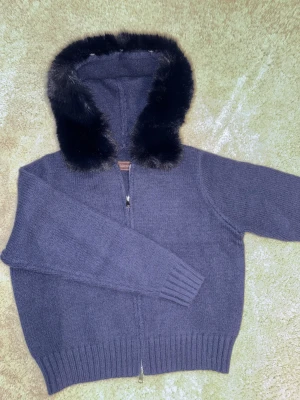 Alessa fur jacket  - Mysig mörkblå stickad hoodie från Aless med fluffig svart päls runt huvan. Tröjan har dragkedja framtill, ribbade muddar och långärmad passform. Perfekt för kalla dagar när du vill vara både varm och snygg.