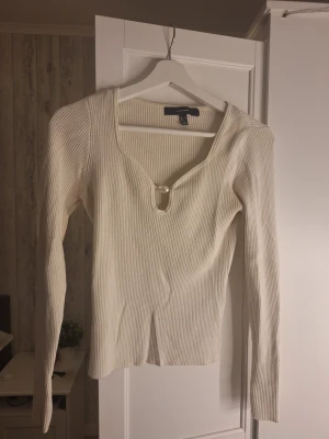 Ribbad vit topp från Vero Moda - Säljer en ribbad vit långärmad topp från Vero Moda i storlek S. Toppen har en snygg v-ringning med metallring-detalj framtill och är figurnära i modellen. Perfekt för dig som gillar stilrena och trendiga plagg med det lilla extra.