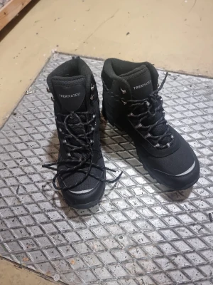 Svarta vinterkängor Trekmates Ice Tech - Svarta kängor från Trekmates med Ice Tech-teknologi, perfekta för vinter. Utrustade med grov sula och metalldubbar för extra grepp på is. Fodrade med mjukt material för värme och komfort. Snörning framtill och robust design, passar dig som vill ha stabilitet och stil.