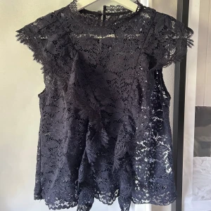 Mörkblå spetsblus med blommönster - Säljer en mörkblå blus i genombruten spets med blommönster. Blusen har korta ärmar och rund halsringning, samt en liten knapp i nacken. Perfekt för dig som gillar romantiska detaljer och vill ha något extra till outfiten.