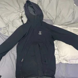 Svart vindjacka från Haglöfs med huva - Svart vindjacka från Haglöfs i storlek S, med justerbar huva och meshfoder på insidan. Jackan har dragkedja framtill och praktiska fickor med dragkedja. Perfekt för dig som gillar outdoor och vill ha en stilren jacka med funktionella detaljer.
