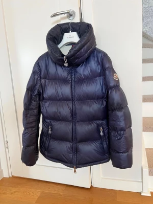 Moncler Douro Giubbotto - Säljer en mörkblå pufferjacka från Moncler med hög krage och två fickor med dragkedja. Man kan ta fram en liten luva ur kragen. Den är storlek 2, skulle säga att den är som en S. Den är köpt på Nk i Göteborg, det finns äkthetsbevis. 💕Den har inget att anmärka på, den är som ny! 