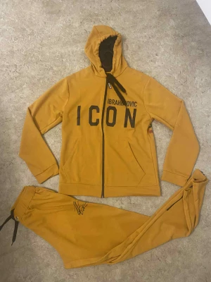 Mustardgul hoodie DSQUARED2 ICON - Säljer en sjukt snygg mustardgul hoodie från DSQUARED2 med svart text 'ICON' och 'IBRAHIMOVIC' på framsidan. Baksidan har en stor print med Zlatan och DSQUARED2-logga. Hoodien har dragkedja, fickor och svart snörning i huvan. Materialet är mjukt och skönt, perfekt för chill.