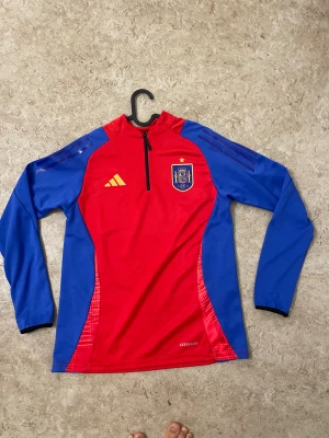 Spaniens långärmad träningströja Adidas - Säljer en röd och blå långärmad träningströja från Adidas med Spaniens landslagsmärke på bröstet. Tröjan har half zip framtill, gula Adidas-loggan och Aeroready-material som andas. Perfekt för fotbollsträning eller chill. Snygg design med kontrastfärgade ärmar.