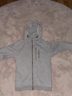 Grå hoodie från Sail Racing barn 150 cm - Snygg grå hoodie från Sail Racing med dragkedja hela vägen och en vertikal zipficka på bröstet. Hoodien har justerbar huva med svarta snören och logga tryckt på bröstet. Perfekt för chill dagar och streetwear. Mjuk och skön bomullsblandning. Storleken är 150 cm