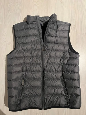 Grå dunväst från Polo Ralph Lauren - Snygg grå dunväst från Polo Ralph Lauren med quiltad design och broderad logga på bröstet. Västen har hög krage, två sidofickor med dragkedja och hel dragkedja framtill. Perfekt för lager-på-lager och chill stil.