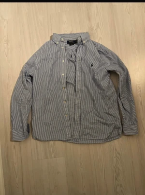 Blårandig skjorta från Polo Ralph Lauren - Snygg blårandig skjorta från Polo Ralph Lauren i slim fit-modell. Skjortan har klassisk krage, knappar framtill och det ikoniska Polo-broderiet på bröstet. Tillverkad i mjuk bomull, perfekt för dig som gillar stilrena och tidlösa plagg.