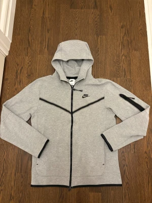 Grå Nike hoodie Tech Fleece - Säljer en äkta Nike Tech Fleece Windrunner Hoodie i färgen Heather Grey med svarta detaljer. Mycket fint skick. Inga fläckar, noppor eller slitage. sömmar är i perfekt skick, endast minimalt slitage på dragkedja. Endast sparsamt använd. Storlek S. Original modell som inte säljs i butik längre. en ganska ikonisk version som Nike inte längre tillverkar