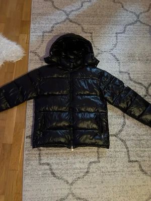 Svart Moncler pufferjacka med huva - Säljer en svart pufferjacka från Moncler med glansig finish och huva. Jackan har dragkedja framtill, två fickor med dragkedja och klassisk Moncler-logga på ärmen. Perfekt för kalla dagar och riktigt snygg streetstil.