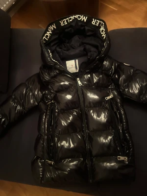 Svart Moncler pufferjacka med huva - Säljer en svart, glansig pufferjacka från Moncler med stor huva och logotyp på ärmen. Perfekt för kalla dagar och riktigt snygg streetstil.