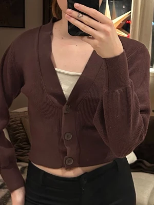 Brun croppad kofta med knappar - Snygg brun croppad kofta med tre stora knappar framtill och v-ringad hals. Tröjan har långa ärmar med ribbade muddar och är stickad i ett mjukt material. Storlek CS, från Gina Tricot