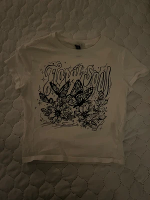 Vit t-shirt med Floral Soul-tryck - Snygg vit t-shirt från H&M med coolt svart tryck som säger 'Floral Soul' och har detaljerade blommor och fjärilar på framsidan. Modellen är kortärmad och har en klassisk rund hals. Perfekt för dig som gillar grafiska prints och vill sticka ut.