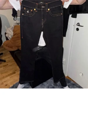 True Religion svarta jeans med kontrastsömmar - Svarta jeans från True Religion med tydliga vita och orange kontrastsömmar, klassiska fem fickor och snygga detaljer på bakfickorna. Rak passform och normal midja, tillverkade i slitstarkt denim. Perfekta för dig som vill ha en clean men ändå unik look.