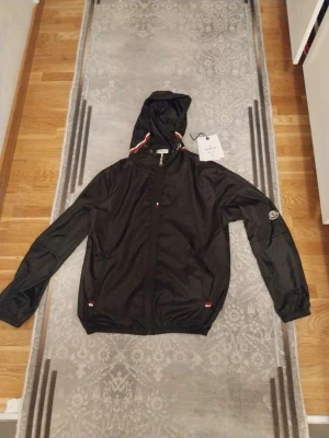 Svart vindjacka från Moncler, M - Svart vindjacka från Moncler med huva och dragkedja framtill. Jackan har Moncler-logga på ärmen och snygga detaljer i rött, vitt och blått vid huvan och dragkedjan. Tillverkad i lätt polyester, perfekt för blåsiga dagar. Klassisk passform och stilren look.