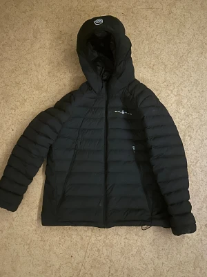 Svart dunjacka från Sail Racing - Svart dunjacka från Sail Racing med huva och dragkedja. Jackan är fylld med certifierat dun (600 fill power) och har en quiltad design. Vind- och vattenavvisande material, andas bra och är fluorkarbonfri. Perfekt för kalla dagar och har två fickor med dragkedja. Den passar dig som är 165-175