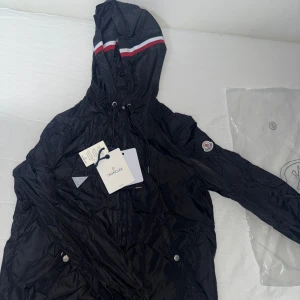 Svart Moncler vindjacka med huva regn jacka  - Svart vindjacka från Moncler med huva och coola rödvita ränder på kanten. Jackan har dragkedja, snörning vid huvan och Moncler-logga på ärmen. Lätt och smidig polyester, perfekt för blåsiga dagar. Snygga silverfärgade knappar på fickorna. Med nfc funkar 