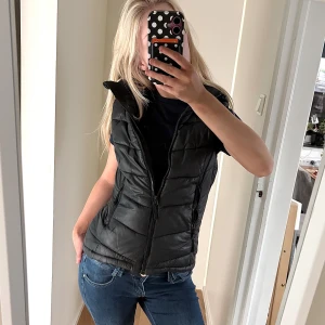 Svart pufferväst från H&M Sport - Svart pufferväst från H&M Sport med hög krage och dragkedja framtill. Västen har två sidofickor med dragkedja och quiltad design. Perfekt att slänga över en hoodie eller t-shirt för en chill look. Materialet är syntet och västen har en normal passform.