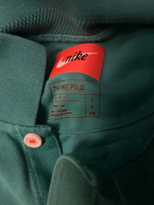 Turkos Nike golf-piké - Vill du vara den snyggaste på golfbanan eller bara en skön vardags piké. Detta är en Nike golf-piké i turkos med korta ärmar och vit knapp framtill. Toppens framsida har små broderade motiv i vitt och mörkblått, bland annat kryss och hand-symboler. Materialet är bomull och modellen är normal passning. Perfekt för dig som gillar sportig och chill stil. 